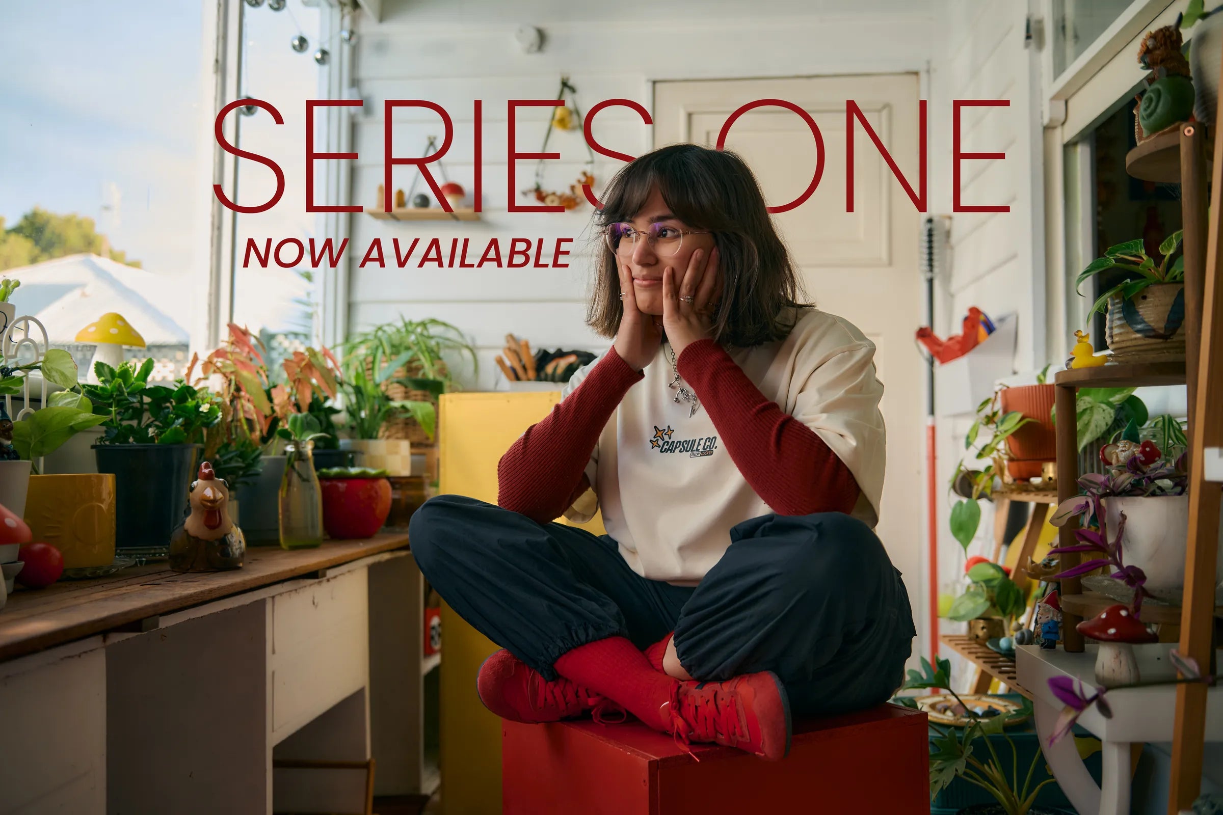 Series One, now available. A model sits cross-legged on a red box in a sunroom full of plants, wearing the Ganbaru Tee in Natural over a red long sleeve.